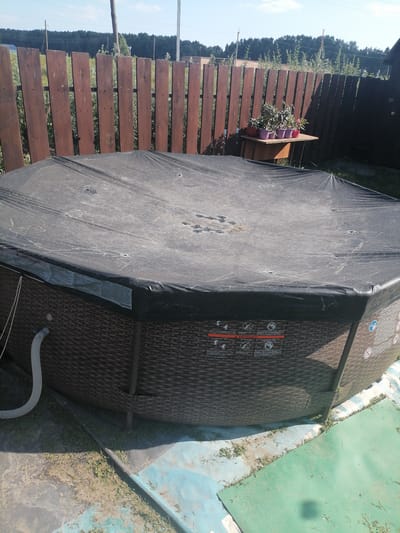 The image shows an outdoor scene in the city of Степной, Russia, on a sunny day. In the foreground, a large, circular inflatable hot tub is covered with a black tarp. The sides of the hot tub are a dark brown wicker-like pattern. A white hose is attached to the side of the hot tub. The hot tub is situated on a patch of ground that appears to be covered by a blue and green tarp or matting, with some grass and dirt visible. In the background, a wooden fence with vertical planks runs horizontally across the frame. Behind the fence, there is a line of green trees, and beyond that, a clear blue sky. On a small wooden table positioned against the fence, several potted plants are visible. The overall impression is of a residential backyard.