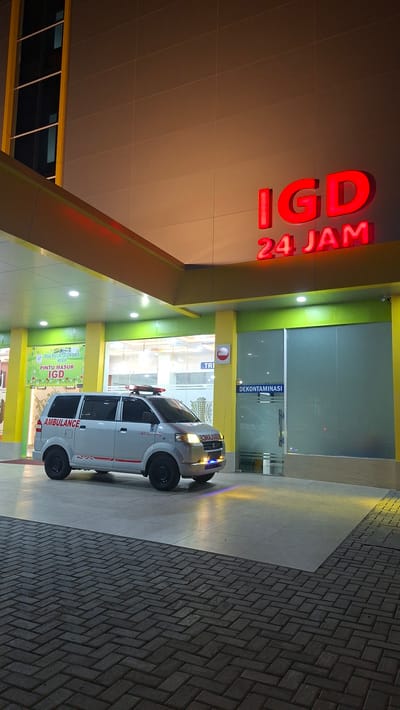 The image shows the exterior of a hospital emergency department (IGD) at night. A white ambulance is parked in front of the entrance, its lights flashing. The building has a prominent red neon sign that reads "IGD 24 JAM," meaning "Emergency Department 24 Hours." The entrance area is lit by yellow and green lights, and the ground is paved with interlocking bricks. Inside the building, through glass doors, more lights and signage are visible, including a sign that says "DEKONTAMINASI" (Decontamination). The scene appears to be in Lampeuneurut Blang, Indonesia. The overall impression is one of readiness and accessibility for medical emergencies.