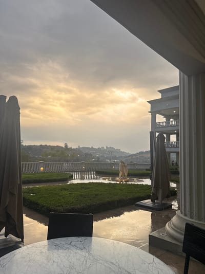 The image depicts an outdoor terrace or courtyard located in Atizapán de Zaragoza, Mexico. The scene is illuminated by the diffuse light of either dawn or dusk, with the sky displaying a mix of grey and warm orange-yellow clouds. The ground surfaces are wet and reflective, indicating recent rainfall.

In the immediate foreground, a round table with a white marble-like pattern is centered, with the back of a dark chair partially visible behind it. To the left, a large, closed, beige outdoor umbrella is partially visible. To the right, a substantial white classical column rises from the lower frame.

The midground features a formal landscaped area bordered by a white balustrade with multiple spherical lights. Within this area, precisely trimmed green hedges form geometric beds. Pathways between the hedges are wet and reflective. A fountain with a light-colored statuary group, depicting multiple figures, is visible centrally in the landscaped section, with water actively present. Another large, closed, beige outdoor umbrella stands to the right within this area.

The background reveals a hilly natural landscape covered with trees, extending towards a distant urban skyline featuring various buildings. On the far right, a classical-style multi-story building with columns and balustraded balconies is visible; two indistinct human figures are discernible on an upper balcony.