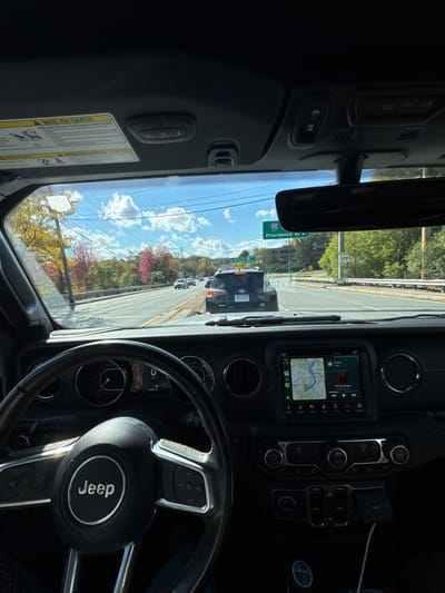 This image captures the interior perspective from a Jeep vehicle, stationary or moving slowly in traffic on a multi-lane road in Newton, Massachusetts, United States. A green highway sign on the right side of the road displays "95 NORTH Providence RI W" and "128 NORTH," confirming the location.
The immediate vehicle ahead is a dark-colored SUV, identifiable by its Massachusetts license plate "6RS 891". The environment features a clear blue sky with scattered white clouds. Trees lining the roadway exhibit vibrant red, orange, and yellow autumn foliage.
Inside the Jeep, the black dashboard, a multifunction steering wheel with the "Jeep" logo, and a central infotainment screen are prominent. The screen displays a navigation map and a music track titled "On My Knees." Below the screen are vehicle controls and a charging cable. A "MISE EN GARDE" warning label is affixed to the sun visor. The scene depicts daytime conditions during the autumn season.