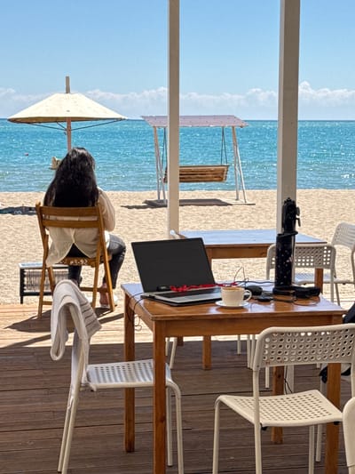 An outdoor scene at a beach cafe on a sunny day. In the foreground, a wooden table is set up with a laptop, a coffee cup, a water bottle, and headphones, suggesting a work or study setup. A white chair with a towel draped over it is positioned at the table. In the background, a person with long dark hair is sitting in a wooden chair, facing away from the camera, looking out at the sea. The beach, the blue ocean, and the sky are visible. The location is Koshkol', Kyrgyzstan.