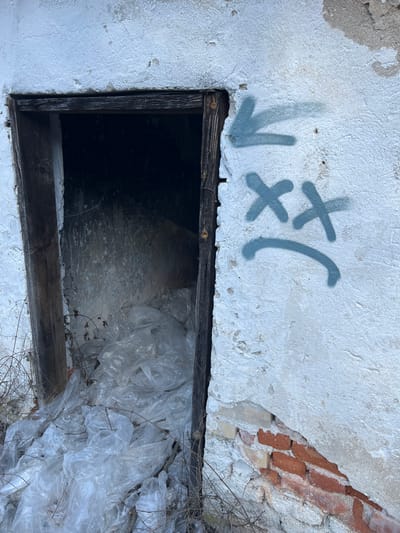 In Stoianovo, Bulgaria, an outdoor scene depicts a derelict structure's doorway. The wall surrounding the entry is covered in weathered, light-colored plaster, heavily cracked and peeling in multiple areas, revealing underlying reddish-brown brickwork at the lower right. A dark, rectangular doorway, framed by aged, dark wooden timbers, lacks a door. The interior visible through this opening is dark, with the floor almost entirely obscured by a large accumulation of crumpled, translucent, clear plastic bags. Dry, sparse vegetation is present at the base of the wall and partially within the doorway. To the right of the doorway, light blue or grey spray paint forms graffiti: an arrow pointing left, an "XX" symbol, and a curved line representing a frowny face. No individuals or specific actions are observed within the frame.
