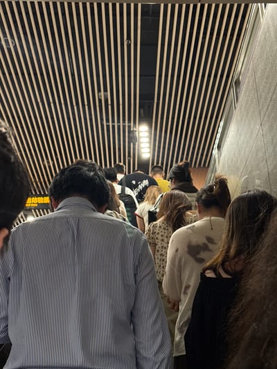 In Xuhui District, China, this image captures a crowd of people ascending an escalator within a modern building. The escalator is adorned with a distinctive ceiling featuring numerous parallel black and beige striped panels. The people in the foreground are mostly seen from the back, indicating a movement upwards. One individual in a blue and white striped shirt is prominently positioned at the front. Other individuals are wearing casual clothing, including a person in a white sweater with dark smudges and another in a black top. The environment appears to be an indoor public space, possibly a transportation hub or a commercial establishment, with a textured grey wall visible on the right side of the frame and soft lighting from overhead fixtures. A yellow sign with Chinese characters and the English text "Exit Gate" is visible on the left, suggesting an exit point.