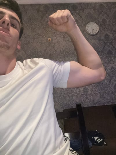 The image depicts an indoor scene located in the city of Durgena, Georgia. A male subject is shown from the chest up, positioned at a low angle and looking directly at the camera. He wears a white, short-sleeved athletic t-shirt and is flexing his right bicep with his hand clenched into a fist. His facial expression is neutral. The background features a wall adorned with dark gray and beige patterned wallpaper, exhibiting a floral or damask motif. A circular analog wall clock, displaying approximately 11:45, is visible on the upper right section of the wall. A small, light-colored square item is positioned on the wallpaper below and slightly to the left of the clock. In the lower right corner, a dark-colored chair or piece of furniture is partially visible. Below this furniture, on a dark surface, a pair of dark athletic shorts with white stripes and another dark item are present. The scene is illuminated by interior lighting.