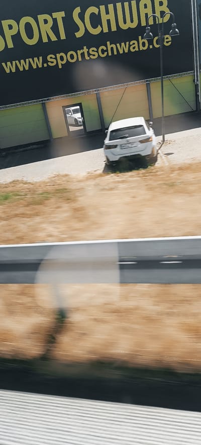 The image depicts a street scene in Winterbach, Germany. In the foreground, a blurry, dry, grassy area is visible, suggesting a roadside or open field. A portion of a modern, grey structure, possibly a building or barrier, runs horizontally across the middle of the frame, with a blurred line marking its edge. Beyond this structure, a paved area is visible, with two white cars. The car on the right is a white station wagon, parked at an angle, showing its rear. A smaller white car is visible through an open doorway of a building in the background, which appears to be a commercial establishment with a dark facade and a yellow and green accent. A large black banner with yellow lettering is prominently displayed above the building, featuring the text "SPORT SCHWAB" and "www.sportschwab.de". A street lamp with two lights is positioned on the right side of the image, casting a shadow. The scene appears to be captured from a moving vehicle, indicated by the motion blur throughout the image.