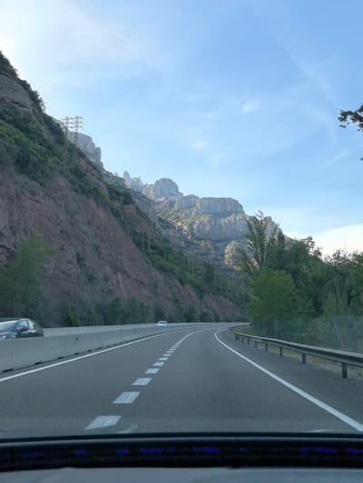 The image presents a view from inside a vehicle driving along a multi-lane highway in a mountainous region, specifically near Collbató, Spain. The road curves gently to the right, featuring white dashed lane markers and a solid white line along the right shoulder. On the left, a rugged and steep mountainside dominates the scene, composed of reddish-brown rock formations interspersed with green foliage. Several electricity pylons and their accompanying power lines traverse the lower slopes of this mountain. Further in the background, the distinctive, jagged peaks of the Montserrat mountain range are clearly visible under a vast, bright blue sky with sparse, wispy clouds. To the right of the highway, a lush green forested area is bordered by a metal guardrail. A sturdy concrete barrier separates the road from the mountain on the left. Three vehicles are captured: a dark car partially visible in the immediate foreground, suggesting the viewpoint from within a car, and two light-colored cars further down the road. The bright, clear sky indicates pleasant weather, likely late morning or early afternoon. The very bottom of the frame shows a portion of the car's dashboard with subtle blue lights. No legible text is visible within the scene.