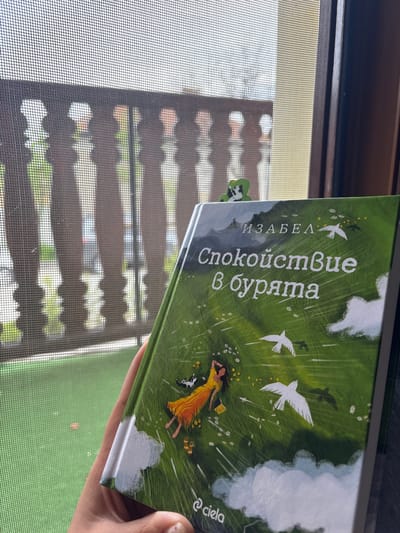 The image shows a book titled "Спокойствие в бурята" (Tranquility in the Storm) by ИЗАБЕЛ (Isabel), published by Ciela, being held in front of a window screen. The book cover features an illustration of a woman in a yellow dress lying in a green field with white birds flying above. A bookmark with a white and black design is clipped to the top of the book. The window screen is visible in the background, revealing a blurry view of what appears to be an exterior environment, possibly including elements of buildings and greenery. The location is not definitively identifiable as Suvorovo, Bulgaria based on visual information alone.