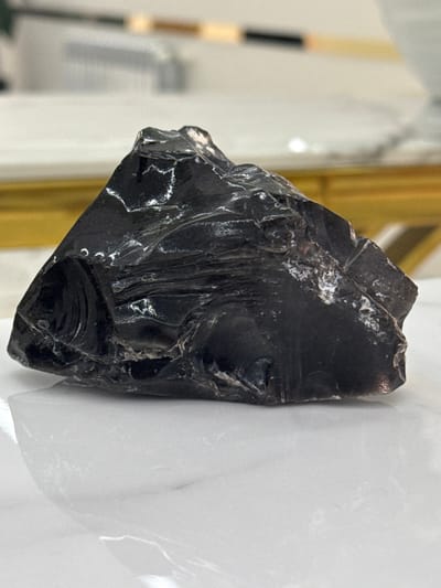 A chunk of black obsidian rock is centrally located. The texture appears glassy with sharp edges and some internal striations or flow patterns. Small white specks are visible on the surface. It is situated near what appears to be plastic wrap or a similar glossy material on the right side. A blurred background includes glimpses of a golden frame and possibly other indeterminate objects. The lighting emphasizes the glossy surface and sharp edges.