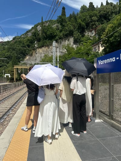 In Gittana, Italy, on a sunny day, a group of people stand on a train platform. The platform is bordered by train tracks on one side and a metal fence and a sign indicating "Varenna - " on the other. In the background, a steep, tree-covered hillside rises, with a few houses nestled into the slope. The people are using umbrellas to shield themselves from the sun. One person in the foreground wears a black t-shirt and black skirt, while others are dressed in white or light-colored clothing. One individual holds a white umbrella, and another holds a black umbrella. The scene captures a moment of waiting at a train station under a bright sky.