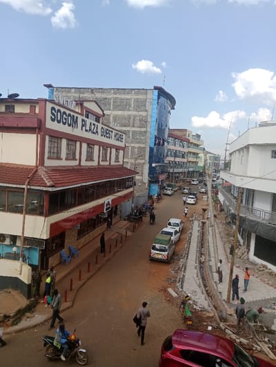 The image shows a street scene in Kapsabet, Kenya, taken from a high vantage point. The focal point is a building on the left side of the image, labelled "SOGOM PLAZA GUEST HOUSE." The building is two-stories with a red roof and a white facade. The building has multiple windows, and there are several people walking in front of it. The street is paved and there are cars parked on both sides. There are several workers laying concrete blocks in the street, seemingly doing road construction. The sky is blue with some white clouds. The buildings on the right side of the image are more modern, with a glass facade, and appear to be part of a shopping center. Several trees are visible in the distance. There are many people walking around, mostly men. The scene is bustling with activity.