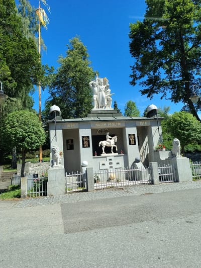 The image depicts a war memorial located in Čkyně, Czechia. The memorial is a grey stone structure with sculptures and inscriptions. At the top of the memorial, there are three white statues of people. Below them, a white statue of a person on horseback is visible. The memorial is flanked by two white lion statues. Inscriptions on the memorial include "1914", "NAŠIM PADLÝM" (To Our Fallen), and "1918". The structure is surrounded by a white metal fence. In front of the memorial, there is a paved area and a road. Trees and foliage are visible in the background, along with a bright blue sky. A tall wooden pole with colorful ribbons is also present to the left of the memorial.