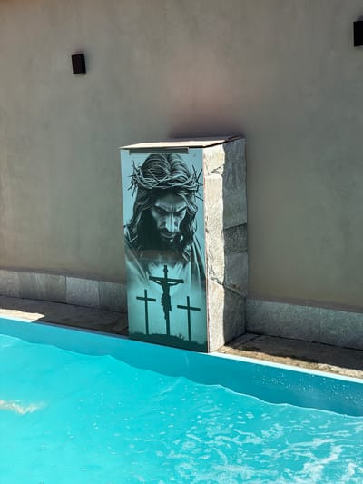 A slightly downward-angled shot shows a wall with a framed black-and-white image of Jesus wearing the crown of thorns, with three crosses behind him. The wall is off-white, and what appears to be a light fixture or sensor is mounted on it. To the right, a stone-like border surrounds the edge of a pool of water, likely in Pirenópolis, Brazil. The pool water is a light blue color.