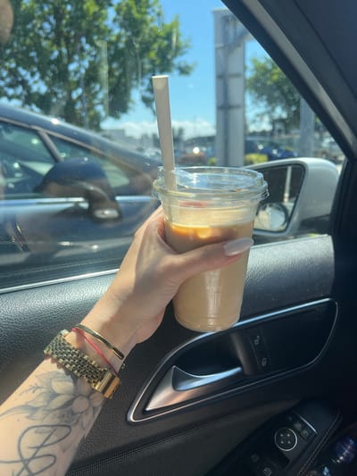 A person's hand, adorned with a gold watch and a red string bracelet, holds a clear plastic cup filled with iced coffee. The cup has a white paper straw and is seen inside a car, with the car's interior visible in the foreground. The person's arm has a tattoo of a flower. Outside the car, through the window, trees and other vehicles are visible under a bright, sunny sky. The location is identified as Heinsberg, Germany.