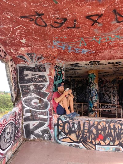 The scene depicts the interior of a derelict, open-sided concrete structure in Kim Long Ward, Vietnam, heavily covered in graffiti. An adult male with a beard, wearing a red t-shirt, dark blue shorts, a backward dark blue baseball cap, and dark blue clogs, is seated on a graffiti-covered concrete ledge. He holds a white smartphone horizontally with both hands, appearing to interact with it, with a white earphone cable connected and hanging. His gaze is directed at the phone's screen.
The structure's interior walls, pillars, and ceiling are extensively covered in various layers of graffiti, including spray paint tags, etched names, and abstract designs in colors such as black, white, red, teal, orange, and grey. The ceiling is predominantly a faded red, densely etched with numerous white inscriptions and overlaid with some black and light blue spray paint. The concrete floor is a reddish-brown hue. Beyond an opening in the structure to the left, blurred green foliage and a light-colored building with a reddish roof are visible under ambient daylight.