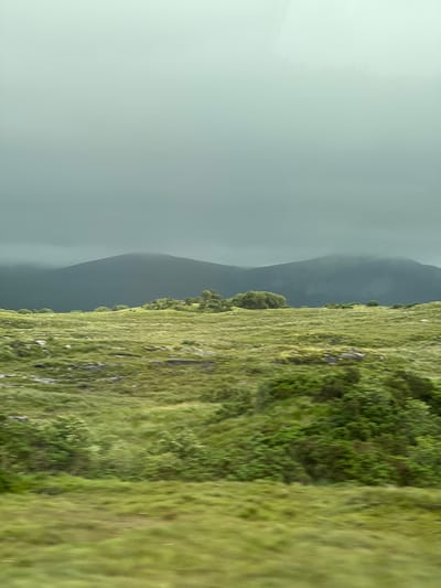 An atmospheric, low-angle shot captures a mountainous landscape under a heavy, overcast sky. The foreground is dominated by rolling green hills covered in grass, with scattered rocks and patches of darker vegetation. In the mid-ground, a line of trees and shrubs adds texture to the undulating terrain. Further in the background, dark, mist-shrouded mountains rise, their peaks obscured by the low-hanging clouds. The scene evokes a sense of quiet and natural beauty, possibly in a rural area like Meenaboy, Ireland, known for its rugged landscapes. There are no people or discernible man-made structures visible in this image.