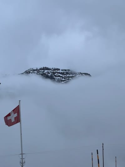 This portrait-oriented image presents a dramatic, snow-dusted mountain peak shrouded in thick clouds or fog, under an overcast sky. The jagged, dark rock face of the mountain rises vertically from the left side of the frame, partially obscured by the white mist, with visible patches of snow highlighting its rugged contours.

In the bottom right corner, a red Swiss flag with a white cross is prominently displayed on a flagpole, gently unfurling, suggesting the presence of wind. To the right, several vertical poles with distinctive black and yellow bands are visible, possibly serving as markers or part of a utility structure, accompanied by thin wires stretching across the frame. A metallic framework, possibly an antenna or part of a cable car system, is also present above the flag.

The scene evokes a sense of high altitude and cold, with the dense cloud cover diffusing the daylight. The overall atmosphere is one of rugged natural grandeur, characteristic of the Alpine environment, and is consistent with the location context of Poschiavo, Switzerland, reinforced by the prominent national flag. No human activity or visible text is apparent in the image.