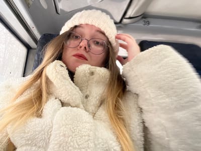 A young adult female subject is depicted in a medium-shot selfie, situated inside what appears to be a passenger vehicle, such as a bus or train, located in the city of Борисово, Russia. The subject has long, straight, blonde hair partially visible beneath a cream-colored knit beanie. She wears round-frame eyeglasses with thin metal rims and has red lipstick applied to her lips. Her attire consists of a thick, textured, cream-colored fleece jacket with button closures. Her left hand is positioned near her head, touching the left side of the beanie. Behind the subject, a dark blue seat back is visible on the right. Above, a light-colored overhead panel displays the number "39". To the subject's left, a window reveals a blurred exterior environment, showing bare tree branches, suggesting movement or a distant, defoliated landscape. The subject is looking directly towards the camera.