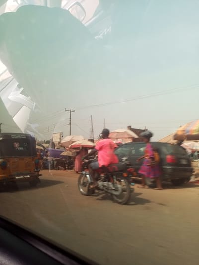 This image presents a vibrant, sunlit street scene, likely captured from the interior of a moving vehicle, evidenced by the reflections of the car's dashboard and windshield in the upper part of the frame. The setting appears to be a bustling market or commercial area in Asokoro, Nigeria.
In the mid-ground, a person wearing a bright pink or red top is seen riding a motorcycle, facing away from the camera. Their left foot is on the ground, suggesting they are either halted or navigating slowly through the street. To the immediate left, the rear of a yellow or orange three-wheeled auto-rickshaw, commonly known as a Keke Napep in Nigeria, is visible. Some indistinct text, possibly "TVC," can be faintly made out on its back. On the right side of the frame, another individual, possibly a woman, is walking or standing near a dark-colored car. She is dressed in a patterned garment with purple, pink, and yellow colors and appears to be carrying an item on her head or in her hands.
The background is filled with numerous market stalls shaded by umbrellas of various colors, indicating a lively trading environment. Power poles and overhead lines are visible against the bright, slightly hazy sky. The general activity depicts a typical busy day with vehicles and pedestrians moving through the street. The bright lighting confirms it is daytime. Due to distance and slight motion blur, specific emotions are not discernible, and aside from possible vehicle branding, no other readable text is clear.