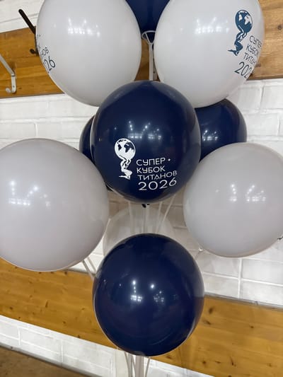 The image shows a group of balloons, some white and some dark blue, arranged indoors against a background of white brickwork and wood paneling. The balloons are clustered together, with some overlapping others. The white balloons have a printed design featuring a figure holding up the Earth along with Cyrillic text which includes the year "2026", with some of the text partially visible on other balloons.