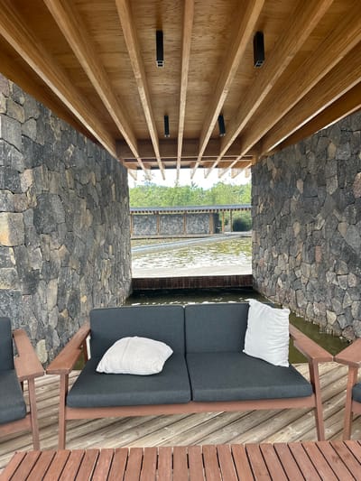 The image captures an outdoor seating area under a wooden-beamed ceiling in Santa Teresa Tiloxtoc, Mexico. A dark gray sofa with two cushions, one white and one light gray, sits on a wooden deck. The sofa is positioned between two walls constructed of rough, gray stones. Beyond the sofa, a body of water is visible, with a wooden walkway leading to another stone-walled structure. Trees and greenery can be seen in the background. The scene appears serene and offers a view of the natural surroundings.