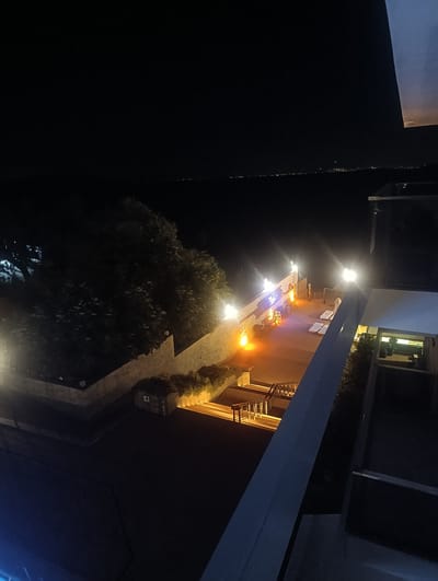 An aerial view captures a dimly lit scene at night, likely in Urla, Turkey, showcasing an outdoor area with illuminated features. A body of water stretches into the distance, dotted with faint lights suggesting a distant city or shoreline. In the foreground, a courtyard area is visible, featuring white lounge chairs, benches, and bright spotlights casting an orange and white glow. A wall with graffiti and plants borders the courtyard. Adjacent to the courtyard, stairs with handrails are illuminated, leading downwards. A large tree with dense foliage stands to the left of the courtyard, partially obscuring the view. The image also shows parts of a modern building structure in the right foreground and middle ground, with illuminated glass elements.