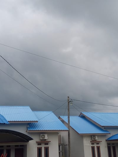 The image displays an overcast daytime scene in Puloblang, Indonesia, featuring multiple buildings, a utility pole, and numerous overhead wires against a gray sky.

On the right side of the frame, several multi-story buildings with light gray or off-white walls are visible. These buildings are topped with bright blue, corrugated metal roofs, some of which extend outwards as awnings. Dark brown, rectangular windows are set into the building walls. At least two outdoor air conditioning units are mounted on the walls; one is positioned between two windows on an upper floor, and another is visible on a lower floor. Faint text, specifically the number "14", can be seen on the wall of one building, and a partial "23" is barely discernible on a lower section.

A wooden utility pole stands vertically on the right side, carrying insulators and a cluster of power lines. Numerous black cables and wires stretch horizontally across the frame, originating from or passing by the utility pole, creating a complex network against the sky.

The left side and upper portion of the image are dominated by a dense, gray, overcast sky, indicating cloudy weather. There is no human activity depicted in the static scene.