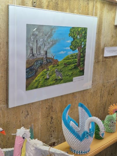 The image captures an art exhibit. A framed painting shows a transition from industrial pollution on the left to nature with trees, animals, and a blue sky on the right, suggesting environmental contrast. To the right of the painting are origami figures, including a swan. The image is set against a textured background. The scene is possibly at or related to the Lake Srebarna nature reserve.