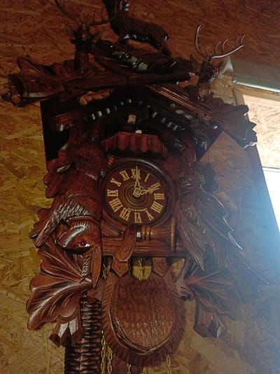 A large, intricately carved dark wooden cuckoo clock is depicted, mounted on a light brown wooden paneled wall in Gorka, Russia. The clock features detailed hunting-themed carvings, including two deer heads with antlers flanking the top, a deer figure positioned centrally above the clock face, and carved birds flanking the sides of the main clock mechanism. A carved rabbit or hare hangs inverted by its legs below the left bird carving, suggesting a hunting trophy. The circular clock face displays gold Roman numerals (I-XII) and two dark hands indicating approximately 02:45. The inscription "MADE IN GERMANY" is visible on the lower portion of the clock face. A small, arched door is situated above the clock face, with a white cuckoo figure partially visible inside. Below the clock face, a carved wooden pendulum, designed as a hunting shield or animal head, is suspended, with two golden chains extending downwards, presumably connected to weights. The scene is illuminated by a strong, unseen light source to the right, creating significant glare on the right side of the clock and the wall. No individuals are present.
