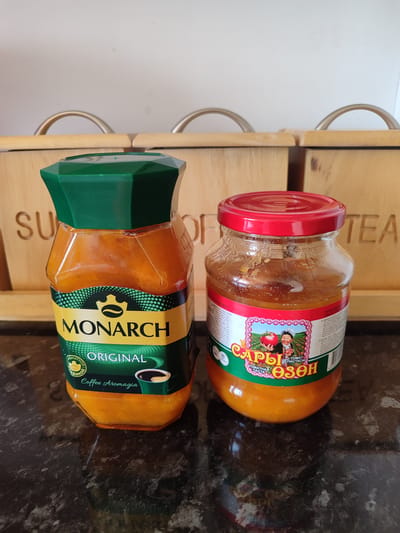 The image displays two jars of jam or spread on a dark, reflective countertop. The jar on the left is tall and hexagonal with a green lid. It has a label that reads "MONARCH ORIGINAL Coffee Aromagia." The jar on the right is shorter and round with a red lid. Its label features a drawing of a man holding a tomato in a field and text that appears to be in Cyrillic, possibly indicating "Tomato Paste." In the background, there are wooden containers with metal handles, and text that reads "SUGAR" and "TEA." The lighting suggests an indoor environment, possibly a kitchen. The image is devoid of any people or specific geographic markers of Batken, Kyrgyzstan.