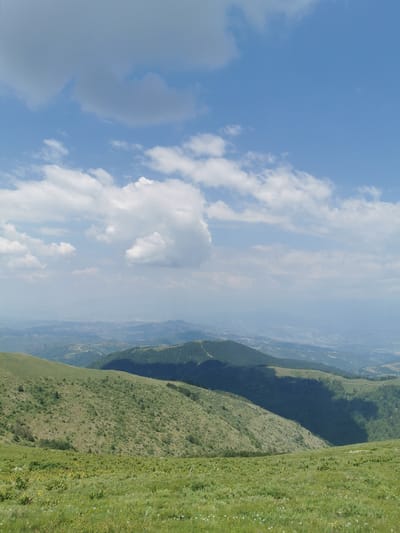 This media file is a landscape photograph taken from a high vantage point, likely a mountain summit or ridge, overlooking a vast, undulating terrain. The scene is characterized by rolling hills covered in lush green grass and scattered vegetation, with a hazy, distant view of more rugged mountains and valleys. The foreground is dominated by a vibrant green meadow, dotted with small white flowers, suggesting a natural, undeveloped environment.

The sky above is a brilliant blue, streaked with white, fluffy cumulus clouds. The sunlight filtering through suggests it is daytime, likely late morning or early afternoon, given the bright illumination and the presence of some shadows cast by the clouds. The overall mood is serene and peaceful, evoking a sense of natural beauty and expansive vistas.

The terrain transitions from the grassy slopes in the foreground to densely forested hills in the mid-ground, and then to more distant, hazy mountain ranges. There appear to be some signs of human presence in the distance, such as faint roads or structures visible on the slopes, but the overall impression is one of a remote and wild natural landscape.

The location is identified as Brestovo, Bulgaria. This context suggests the photograph captures the characteristic mountainous scenery of the region. There is no visible text in the image.