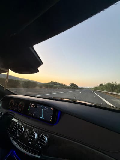 The image captures the interior of a luxury car, likely a Mercedes-Benz, from the perspective of the driver or front passenger. The car is in motion, driving on a highway during sunset or sunrise. The dashboard is clearly visible, featuring a digital instrument cluster and a large infotainment screen displaying a navigation map. The map indicates the location in Greece, showing the city of Arta as a destination or a point on the route. The car's interior is illuminated with ambient blue lighting. The road ahead is open with clear lanes, and the surrounding landscape consists of vegetation and hills, bathed in the warm hues of the setting or rising sun. The sky transitions from a pale orange at the horizon to a light blue. There are no people visible in the frame.