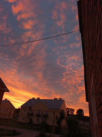 This media file captures a vibrant sunset over a residential area in Medvedyovka, Russia. The sky is a dramatic canvas of orange, pink, and deep blue hues, with scattered clouds catching the last rays of the sun. The setting is characterized by multi-story apartment buildings, their yellow facades contrasting with the warm sky.

In the foreground, a portion of a brick building occupies the right side of the frame, its textured surface adding a sense of depth. To the left and center, two apartment buildings are visible, their roofs silhouetted against the colorful sky. The architecture suggests a relatively modern, yet functional, urban or suburban environment. Bare trees and some foliage dot the landscape between the buildings and further into the background, where a gentle incline suggests rolling terrain.

The time of day is clearly sunset, indicated by the intense colors in the sky and the lengthening shadows. The weather appears calm, with the dramatic cloud formations being a product of the sunset itself rather than a storm. The scene evokes a sense of peaceful transition from day to night, with the warm colors of the sky lending a touch of natural beauty and tranquility to the man-made structures. There is no visible text in the media file.