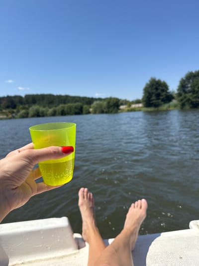 In Tiutky, Ukraine, on a bright, sunny day, a person is relaxing on a boat on a calm body of water. Their bare feet are visible, propped up on the edge of the boat. In their hand, they hold a neon yellow plastic cup filled with a bubbly, light-colored beverage. The background features a dense line of green trees bordering the water and a clear blue sky. The scene suggests a peaceful outdoor recreational activity.