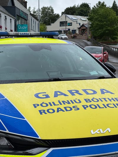 In Donegal Town, Ireland, a Garda Síochána (Irish police) vehicle is parked on a wet road. The car is yellow with blue accents and has the word "GARDA" emblazoned on the light bar and the hood, along with "POILINIÚ BOITHRE" and "ADS POLICE" in blue lettering. The vehicle's hood is covered in raindrops. In the background, a building with a sign reading "Quay Restaurant & Bar" is visible. A red car and a white car are also parked on the street. The sky is overcast.