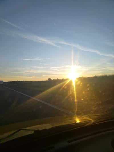 The image depicts a roadside view of a sunset, likely on a highway, with the sun prominently featured. The sky is a gradient of blue, transitioning to yellow and orange near the horizon, with thin wispy clouds. The sun, intensely bright, casts strong lens flares and light rays across the scene. In the foreground, a dark, reflective surface, presumably a car's dashboard or windshield, is visible, showing reflections of the sunlight. To the left, a highway stretches into the distance, with a white dashed line marking its edge. A portion of a white truck with a logo that appears to be "DPD" is visible on the left side of the highway. The background consists of a grassy embankment or field with scattered trees and vegetation. There are no people clearly visible in the image. The overall impression is one of a journey during a sunset. Without further information, the specific location in Szczawin, Poland, cannot be confirmed from the visual evidence alone.