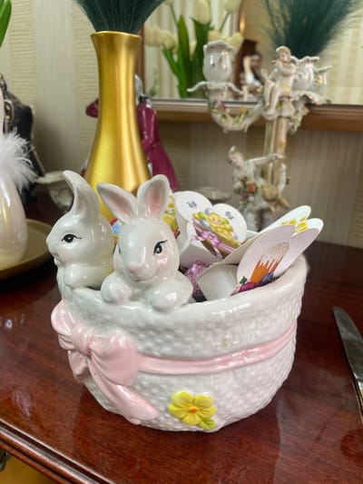 A close-up shot shows a ceramic bunny figurine on a dark wooden surface. The figurine is white with two rabbits near the base. A pink ribbon-like design encircles the container. There is a yellow flower with green leaves on the side. The container appears to be filled with small cards or decorations. In the background, a mirror reflects a decorative gold object with a green feather and a figurine with cherubs. A knife is partially visible in the upper right corner of the frame. The location is not discernible.