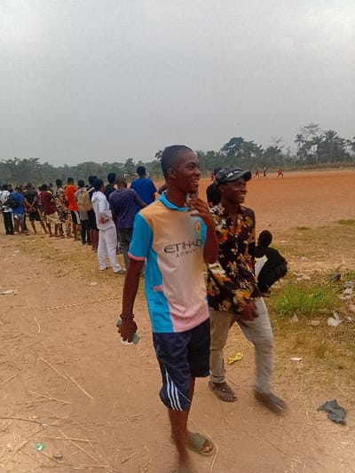 A group of people is standing outside on a sandy ground. The group is predominantly composed of younger people, possibly students. Two young men are prominent in the foreground. One is wearing a colorful shirt and dark shorts, holding something small in his hand. Another individual, wearing a dark floral shirt and light pants, is standing behind him. The backdrop shows a line of trees. The scene suggests an outdoor gathering or activity.