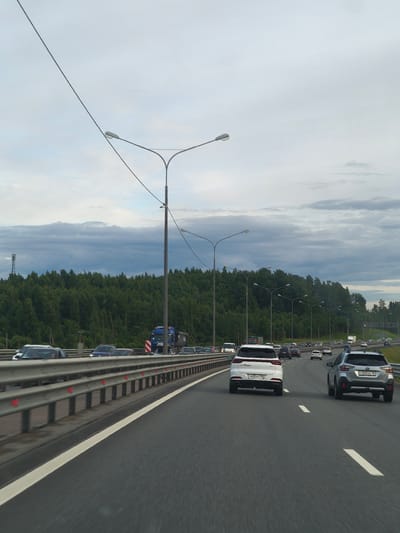 The image shows a highway with multiple lanes of traffic moving under an overcast sky. A dense forest lines the right side of the highway, with a telecommunication tower visible in the distance. Several cars and a truck are on the highway. The location is identified as Korabsel'ki, Russia. The scene appears to be a typical daytime highway view.