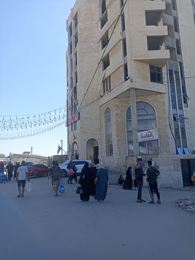 The image depicts a street scene in the city of Gaza. In the foreground, several people are walking and interacting. A man in a white t-shirt and shorts carries a plastic bag. Another man with a backpack walks past a red car. A group of women, some wearing hijabs, are seen walking and conversing. The background features a large, multi-story building constructed with light-colored stone blocks. Some parts of the building appear unfinished, with exposed windows and structural elements. A sign with Arabic script is visible on the facade of the building. Overhead, decorative string lights are strung across the street. The sky is clear and blue. The overall environment suggests a daytime scene in an urban area.