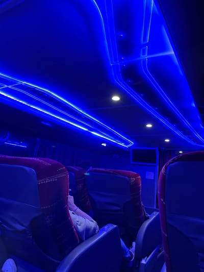 The image depicts the interior of a bus, likely at night, illuminated by vibrant blue LED lighting along the walls and ceiling. Seats with patterned red and blue upholstery line the aisle, occupied by at least one passenger. Small spotlights accent the wall features. The overall ambiance is modern and futuristic due to the intense blue lighting.
