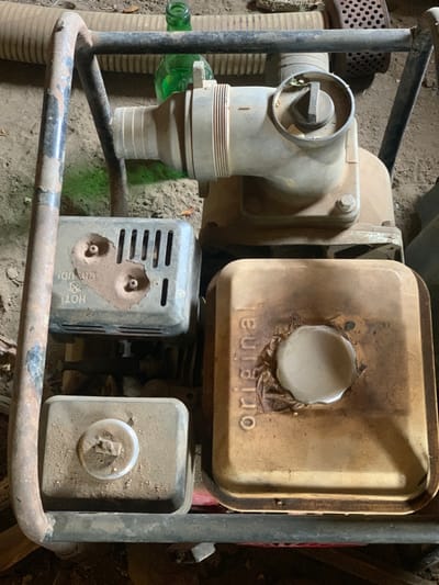 The image shows a water pump in a dirt and rock-filled area. The pump is in working order, with the metal tubing attached to the engine. The engine has a rectangular air filter with the word "original" visible. The engine is mounted on a black metal frame that is attached to a metal pipe. The ground surrounding the pump is dry and dusty. The location of the pump is in Egbeda, Nigeria (latitude: 7.12, longitude: 5.362). There are no people or animals visible in the image.