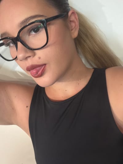 A young woman with blonde hair tied in a ponytail and wearing black-rimmed glasses is shown from the chest up. She is wearing a black tank top. Her tongue is sticking out. The background is a plain white wall. The image appears to be a selfie taken indoors.