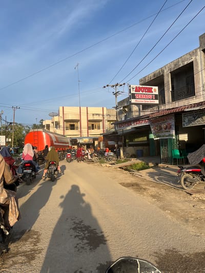 The image depicts a street scene in Alurcucur, Indonesia, likely during the late afternoon due to the long shadows. The street is paved with asphalt and shows signs of wear and tear, with some patchy areas. A prominent red and silver tanker truck is stationary on the road, partially obstructing the view of the buildings lining the street.
Several people are on motorcycles, suggesting traffic or congestion. Some riders are visible, with a few passengers, including women wearing hijabs. The buildings are a mix of styles, with a more modern, multi-story structure on the right featuring balconies and concrete construction. A sign on this building reads "APOTEK TERPADU 1" with smaller text below. Further down the street, another building appears to be a shop or warung with a sign depicting food items.
The environment includes utility poles with overhead wires, a satellite dish on one of the buildings, and vegetation growing along the side of the road, some appearing somewhat overgrown. The sky is clear blue with wispy clouds. The overall impression is of a busy, everyday street in a developing urban or semi-urban area.