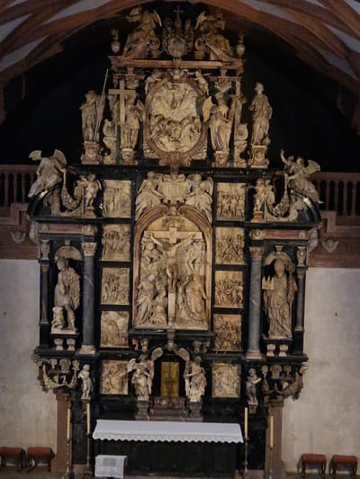 An elaborate, multi-tiered altarpiece, identified as a prominent feature within the St. Peter and Alexander Collegiate Church in Aschaffenburg, Germany, occupies the central frame. The altarpiece is composed of dark, polished stone or wood structural elements framing numerous lighter, carved limestone or alabaster figures and relief panels.
The structure is architecturally complex, featuring columns, arched niches, and pedestals across multiple vertical and horizontal sections. The central focal point is a large crucifixion scene, with Christ on the cross flanked by figures at its base, set within an arched recess. This central element is surrounded by multiple rectangular and circular relief panels depicting detailed biblical narratives with numerous figures in dynamic poses. Large, standing figures, likely saints, are positioned within niches to the left and right of the central crucifixion.
Higher tiers feature additional standing figures, including numerous winged angels and putti, integrated into decorative elements and further relief compositions, culminating in a richly sculpted crowning element at the very top.
At the base of the altarpiece, a dark altar table is covered with a white, scalloped cloth. Two slender, white candles in metal holders flank the altar. On the floor in front, two pairs of dark red, upholstered stools are positioned to the left and right. The surrounding environment consists of light-colored walls in the lower sections, transitioning to a darker background behind the upper altarpiece. A dark wooden balustrade, indicative of an upper gallery, is partially visible on both sides in the background, beneath a dark, possibly coffered or beamed, arched ceiling. No individuals are present in the immediate vicinity of the altar.