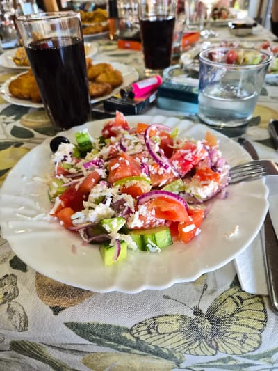 The image shows a plate of Bulgarian salad (shopska salad) on a table covered with a light-colored cloth patterned with butterfly motifs. The salad contains chopped tomatoes, cucumbers, red onion, and crumbled white cheese. A fork is partially visible in the salad. Nearby are glasses and other food items, possibly part of the same meal. The location is Stroevo, Bulgaria.