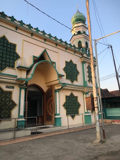 The image shows the exterior of a mosque with a green dome and minaret, located in the city of Keboguyang, Indonesia. The mosque has a white facade with green and gold accents, featuring star-shaped windows with intricate green lattice work. An arched entrance with a wooden door is visible on the right side of the building. A sign above the entrance indicates "TEMPAT WUDLU PRIA," meaning "Men's Ablution Area." The mosque is situated next to a street, with utility poles and wires in the foreground. In the background, a traditional Indonesian house with a tiled roof can be seen. The sky is clear and blue, suggesting it is daytime.