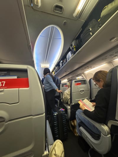 The image depicts the interior of a commercial aircraft cabin, likely a Boeing 737, with passengers either boarding or deplaning. The specific location in Arlington, United States, is not discernible from the visual evidence.
In the foreground, the lower portion of a light-colored sneaker belonging to the photographer is visible at the bottom left. To the right, a female passenger with light brown hair and eyeglasses is seated, facing left, wearing a dark long-sleeved top and blue jeans. She is engrossed in reading a book, holding a pink ruler to mark her place. Under her seat, a brown and tan tote bag is partially visible.
In the center of the image, another female passenger with dark hair, wearing a light blue long-sleeved top and dark pants, stands in the narrow aisle. She is holding the handle of a dark, hard-shell rolling suitcase adorned with colorful tags or decorations. Her stance suggests she is waiting to move forward or find her seat.
Further down the aisle, the back of a third passenger's head is visible, wearing headphones and a light-colored jacket, seated in an aisle seat. The seatbacks in the rows ahead display red labels with "737" printed prominently, along with "REVISION."
The overhead bins are visible on both sides of the aisle, many of them closed and appearing full of carry-on luggage, primarily dark-colored bags. A bright blue accent light illuminates the ceiling near an "EXIT" sign with an arrow pointing left, located on the ceiling panel above the aisle. The cabin features standard artificial overhead lighting. The floor is covered with dark carpet. The overall scene shows a typical passenger environment within an airplane.