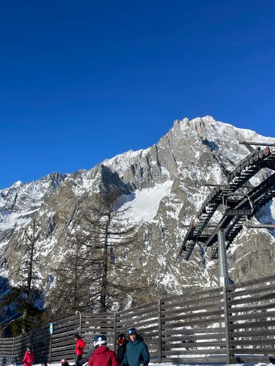 This image depicts a sunny winter day in a mountainous ski resort in Courmayeur, Italy. The scene is dominated by a vast, clear blue sky on the left, contrasting with rugged, snow-covered mountains that fill the right and central parts of the frame.

In the foreground, on the right side, several people dressed in winter jackets and helmets are visible. They appear to be standing or waiting near a tall wooden fence or barrier, suggesting they are at a ski lift station or a viewing area. Above them and extending across the upper-mid section of the image is a large, dark structure of a ski lift, possibly a gondola or chairlift mechanism, with its operational components clearly visible.

The mid-ground features a steep, rocky mountain slope partially covered in snow. Sparse, bare-branched trees, typical of an alpine winter landscape, are scattered across this section, adding texture. A prominent patch of smooth, white snow stands out against the darker rocks of the mountain.

The background reveals more snow-dusted mountain peaks under the bright, cloudless sky. The crisp light indicates a clear day, likely in the morning or early afternoon, with perfect conditions for winter sports. The overall atmosphere is one of serene natural beauty combined with the activity of a ski resort. No discernible text is visible.