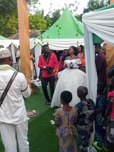 This image captures an outdoor wedding celebration in Amokvo, Nigeria. The scene is set on artificial green turf, beneath white draped canopies and a prominent green peaked tent adorned with artificial green leaves.
In the center, a woman identified as the bride and a man identified as the groom are the focal subjects. The bride wears an off-the-shoulder white ball gown with silver embellishments, a prominent necklace, and white fingerless gloves, holding a silver ceremonial object in her right hand. Her dark hair is styled back. The groom, positioned to her left, wears a red tuxedo jacket with black lapels, a black shirt, black trousers, and dark dress shoes, holding a dark object in his left hand. Both look forward, slightly to their right.
In the immediate foreground, a man in a white long-sleeved top and pants, wearing a white cap and a "GUCCI" branded strap across his back, faces away from the camera, holding a smartphone as if recording. Several children are also visible in the foreground, facing away, observing the scene. One girl wears a leopard-print and floral peplum dress, and a boy wears a blue patterned outfit.
On the artificial turf, two circular woven mats are visible, each holding several Nigerian Naira banknotes (green, yellow, and red denominations). Numerous guests are present in the background, many wearing traditional vibrant African attire or semi-formal clothing. A man in a maroon traditional outfit stands to the bride's right. The background also features natural foliage, indicating an open-air setting.