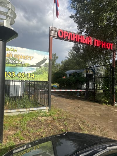 The image captures the entrance to a facility with a prominent red neon sign that reads "Орлиный приют" (Eagle Shelter). The sign is mounted on a wooden frame supported by metal posts. To the left of the entrance, a large banner displays an image of an eagle in flight over a body of water and lush greenery, along with the text "ДЫХА" and a phone number "103-95-34". A black metal fence borders the property, and a white barrier with red stripes is partially lowered across the dirt road leading into the facility. In the background, a small, dilapidated building with a metal roof is visible, surrounded by trees and bushes. The ground is muddy, indicating recent rain. A Russian flag is seen flying from a flagpole above the neon sign. The scene appears to be located in the city of Melikhovskaya, Russia.